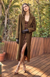 A woman is wearing an elegant, long brown coat with a double-breasted design and wide lapels. Underneath, she has on a brown dress with a slit that reveals her leg. She is also wearing black gloves and brown high-heeled shoes with ankle straps. Accessories include a gold necklace and hoop earrings. She has shoulder-length brown hair and is standing on a wooden deck outdoors, surrounded by green foliage.